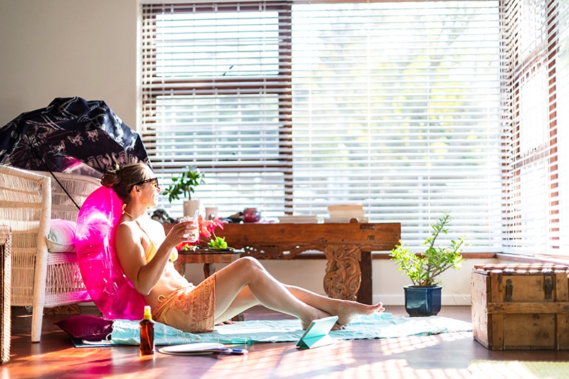 Woman wearing swimsuit and sunglasses sitting on towel and innertube inside on the living room floor. In front of a sunny window trying to stay cool.