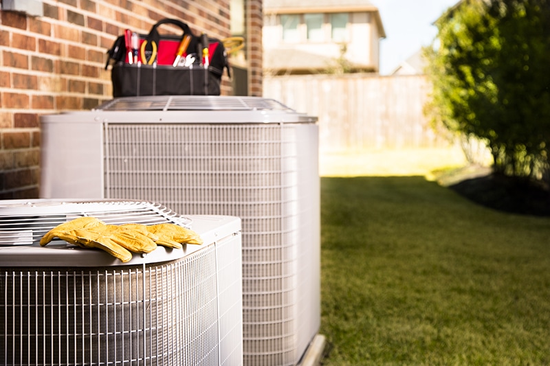 Bag of repairman's work tools, gloves on top of air conditioner units outside a brick home. Service industry, working class.